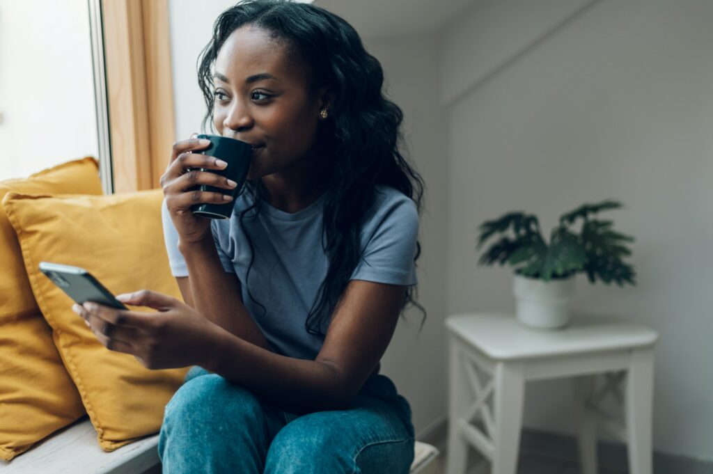 African american woman sitting on windowsill with a cup of coffee and a smartphone
